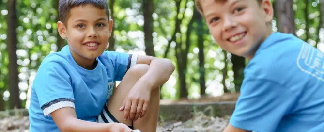 Twee jongens in blauwe T-shirts zitten lachend op een stenen trap in een zonnig bos. Een van de jongens houdt een stuk komkommer vast en kijkt direct in de camera, terwijl de andere jongen rechts van hem ook vrolijk glimlacht. De achtergrond bestaat uit een dicht bladerdek van groene bomen.